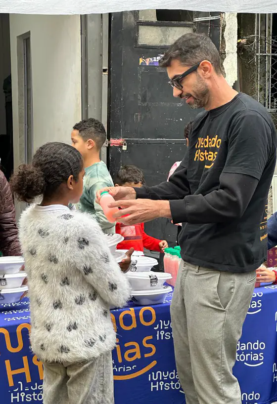 Voluntário de moletom da Mudando Histórias entrega uma bebida para uma criança na mesa de distribuição