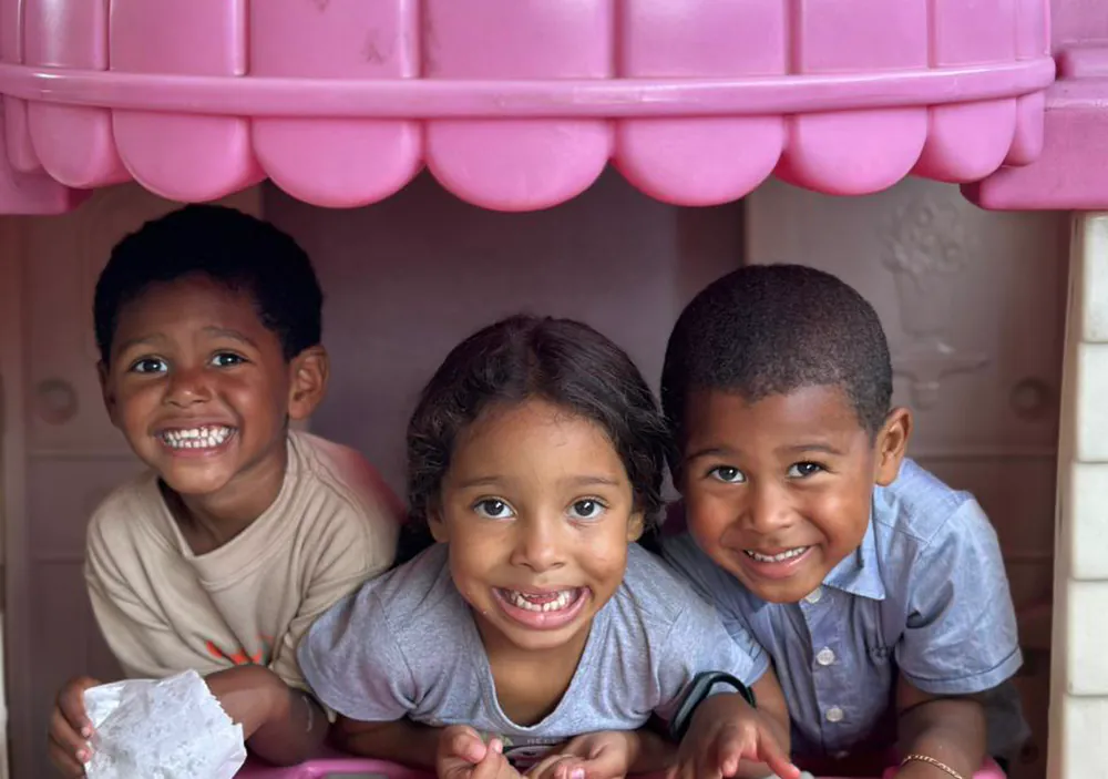 Três crianças sorriem debruçadas na janela de uma casinha de playground