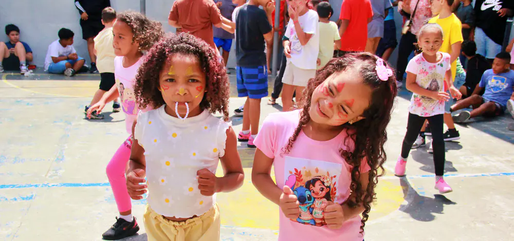 Duas meninas com o rosto pintado posam para a foto em um pátio cheio de crianças brincando