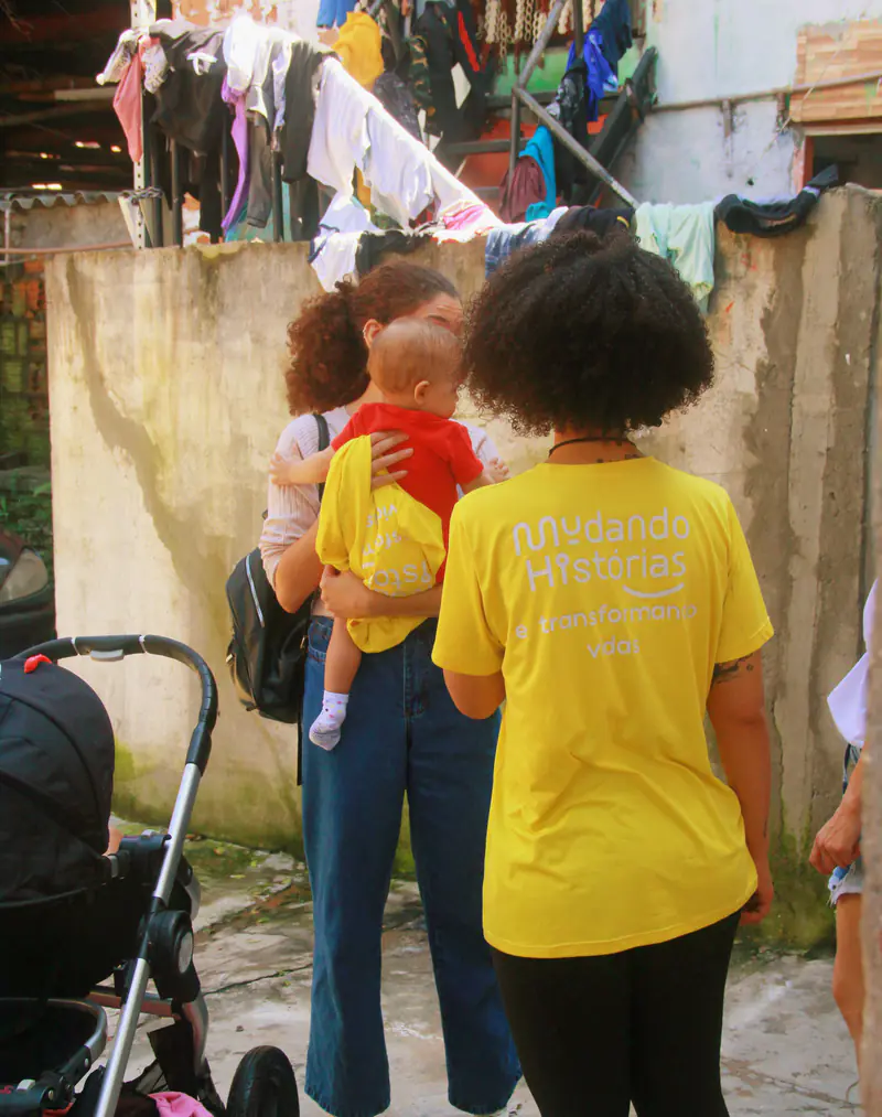 Voluntária de camiseta amarela da Mudando Histórias ao lado de uma mãe que carrega um bebê em um beco com roupas no varal
